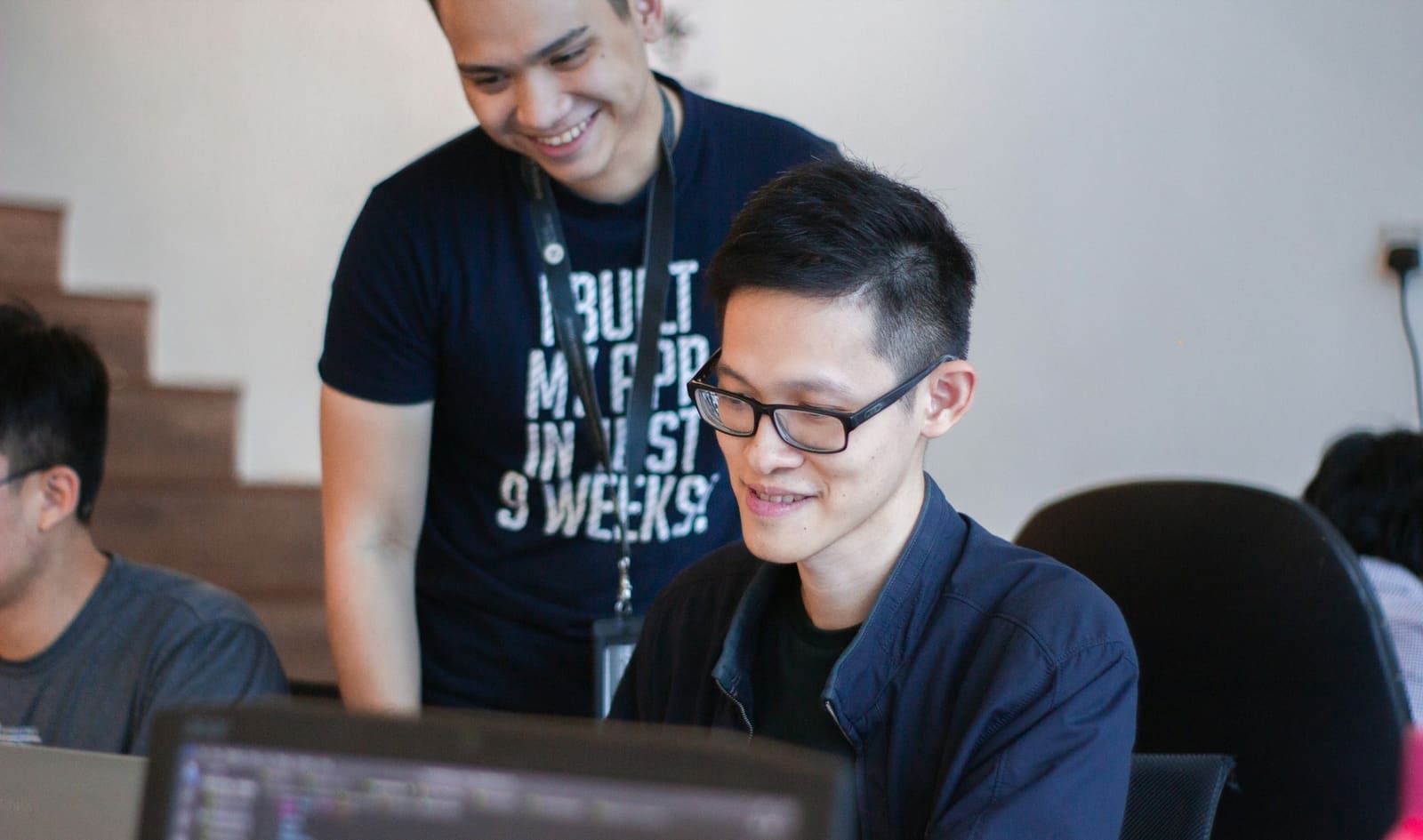 Two developers at a laptop, one standing and smiling as he watches the other work.